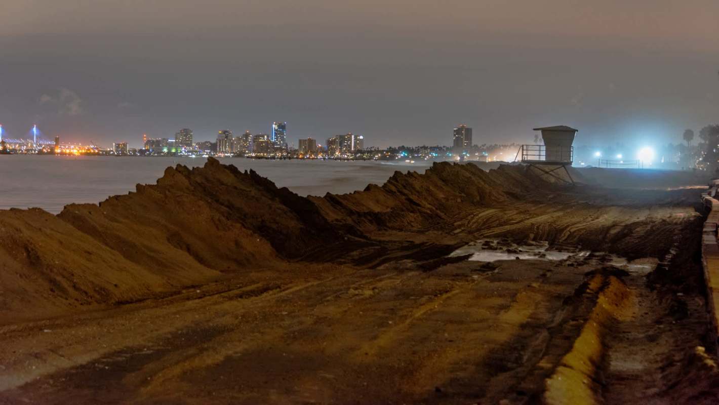 A sandy beach at night with city lights and a lifeguard tower in the distance.