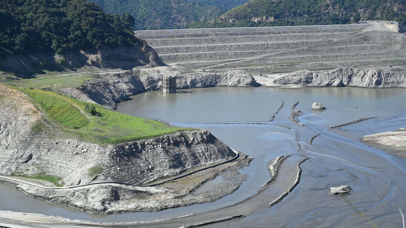 A dry reservoir with exposed banks, water sections, and surrounding green hills.