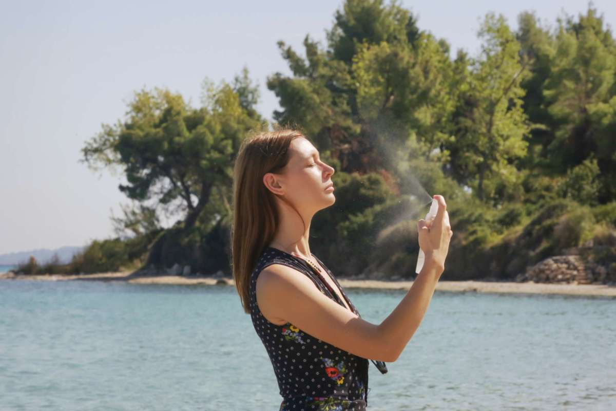 A woman sprays herself with a mist by a beach surrounded by trees.