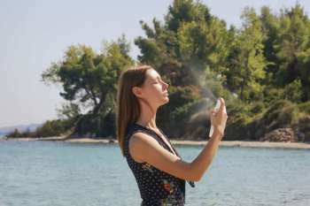 A woman sprays herself with a mist by a beach surrounded by trees.