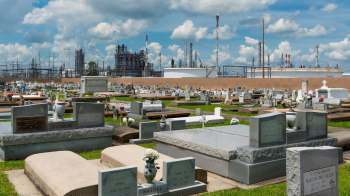 A cemetery with gravestones and flowers, overlooked by petrochemical plants and a cloudy sky.