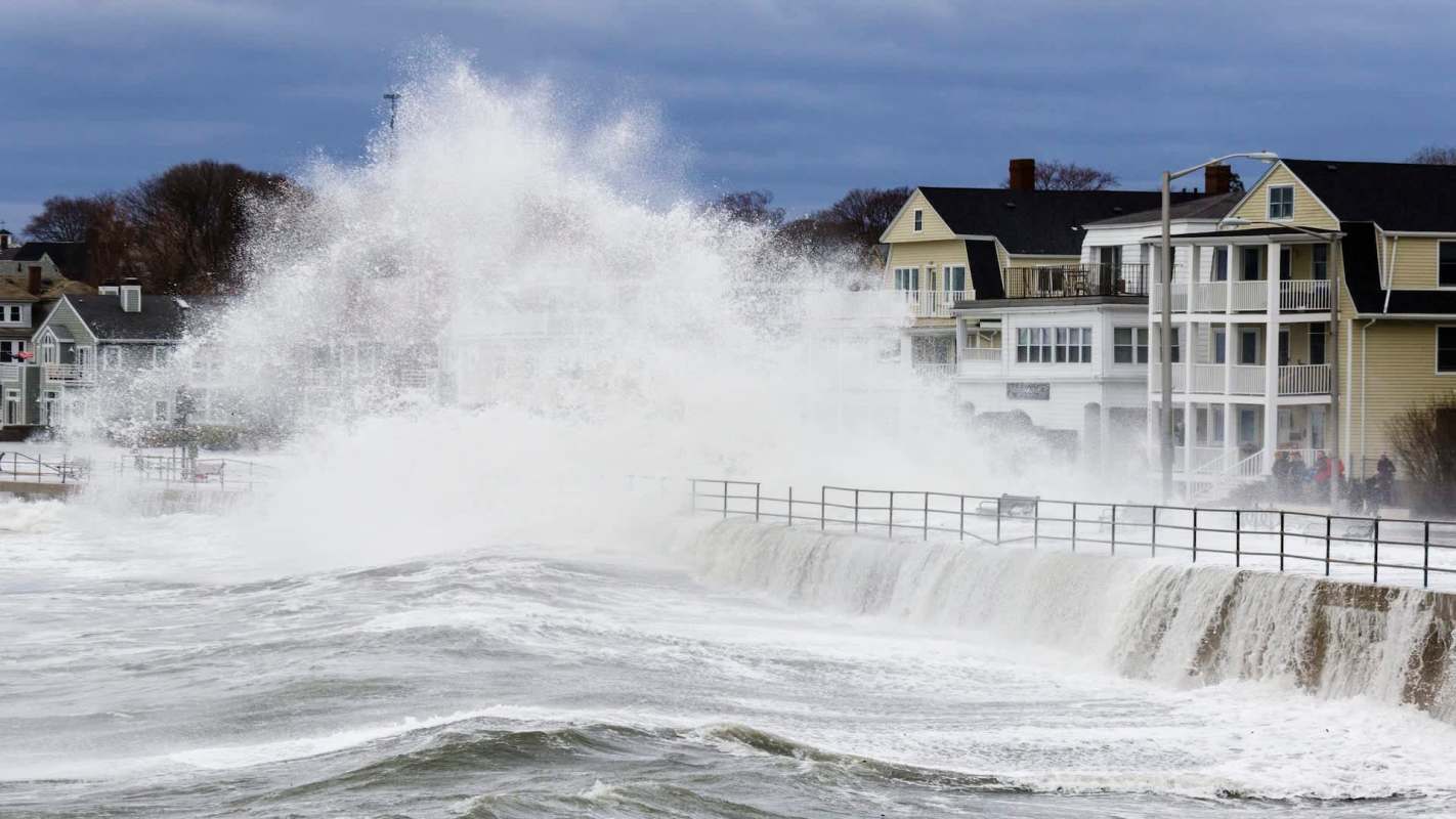 Waves crash over a seawall near houses along a stormy coastline.