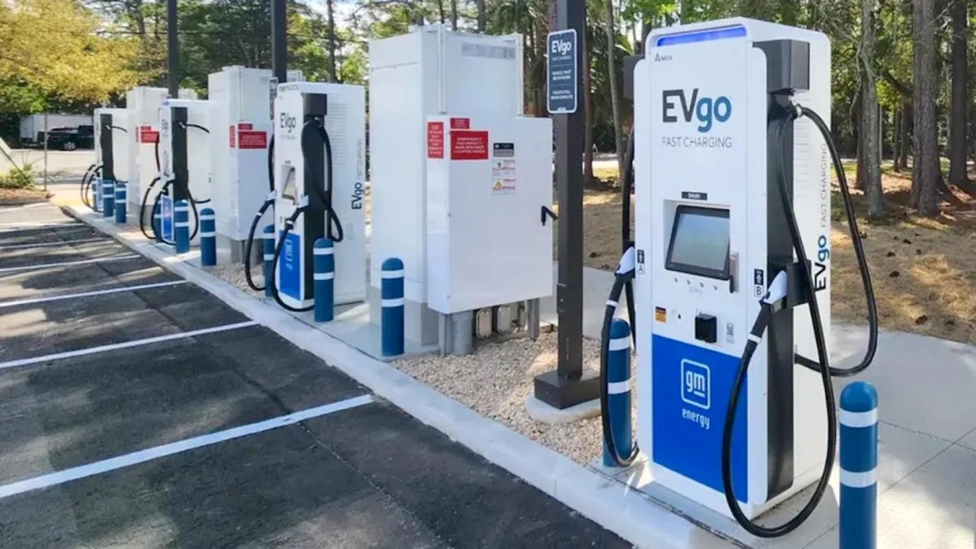 Four EVgo fast charging stations with charging cables in a parking lot surrounded by trees.