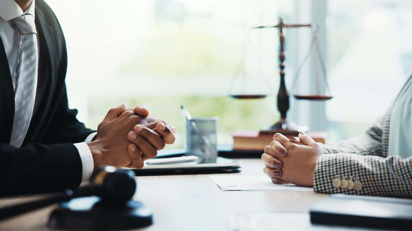 Two people sit at a table with clasped hands, a gavel, and a scale of justice in the background.