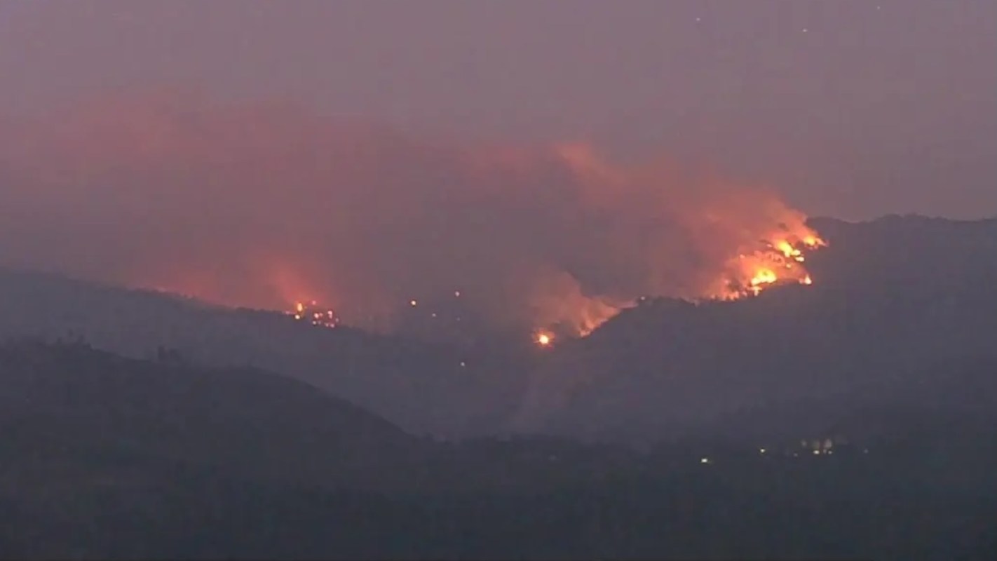 A wildfire burns on a mountain, sending flames and smoke into the sky at dusk.