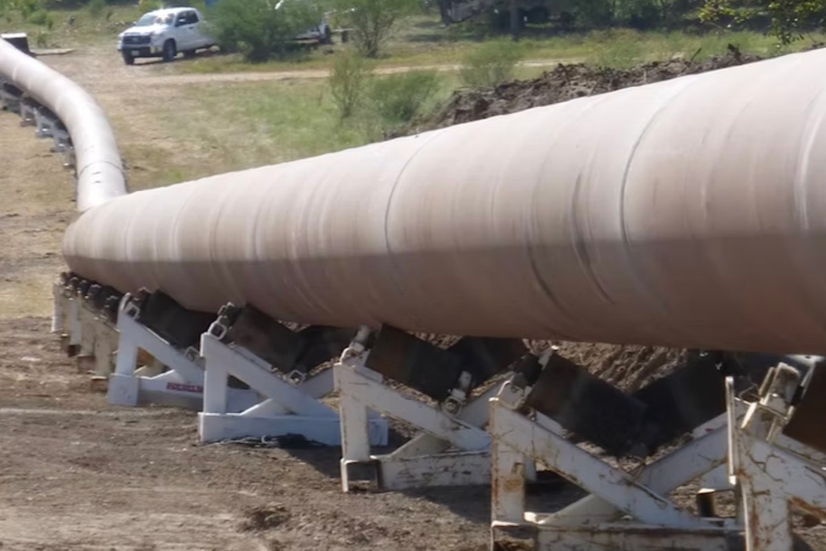 A long pipeline supported by white brackets stretches across a dirt landscape.
