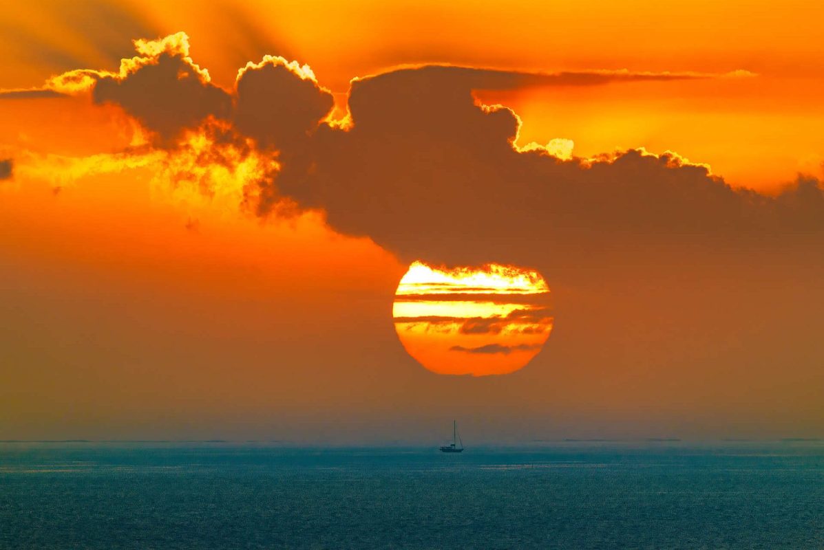 A sunset over the ocean with clouds and a small sailboat in the foreground.