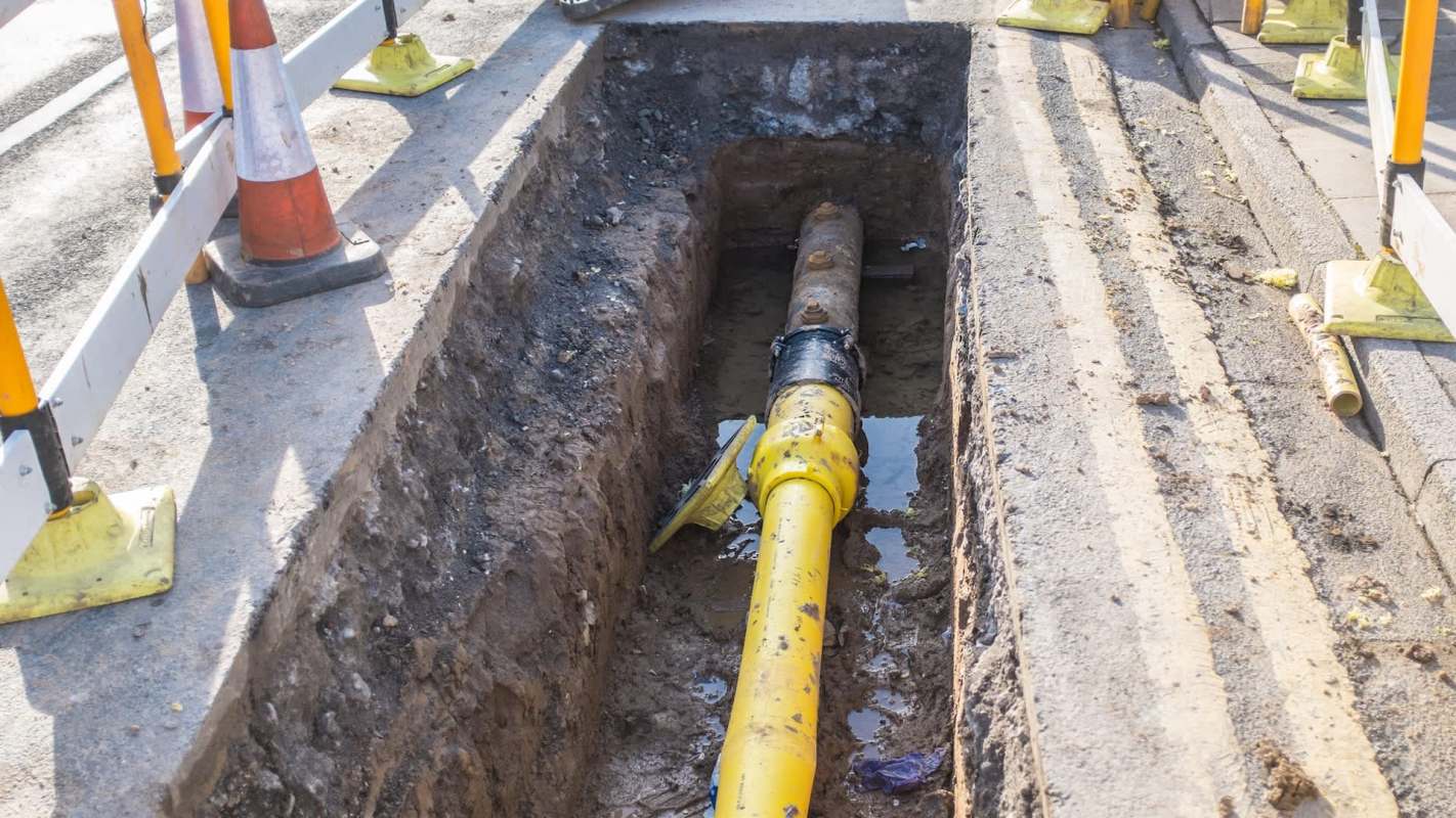 A trench containing a yellow pipe, surrounded by construction barriers and visible water accumulation.
