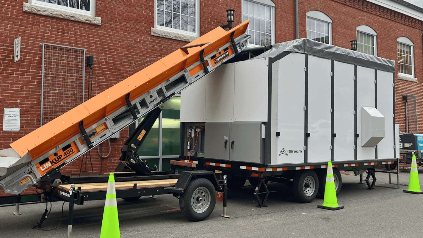 A mobile unit with an orange conveyor ramp parked next to a brick building, surrounded by traffic cones.