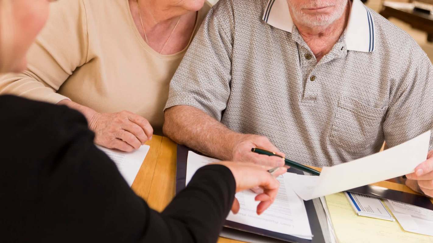 An elderly couple sits at a table reviewing documents with a person pointing at a paper.