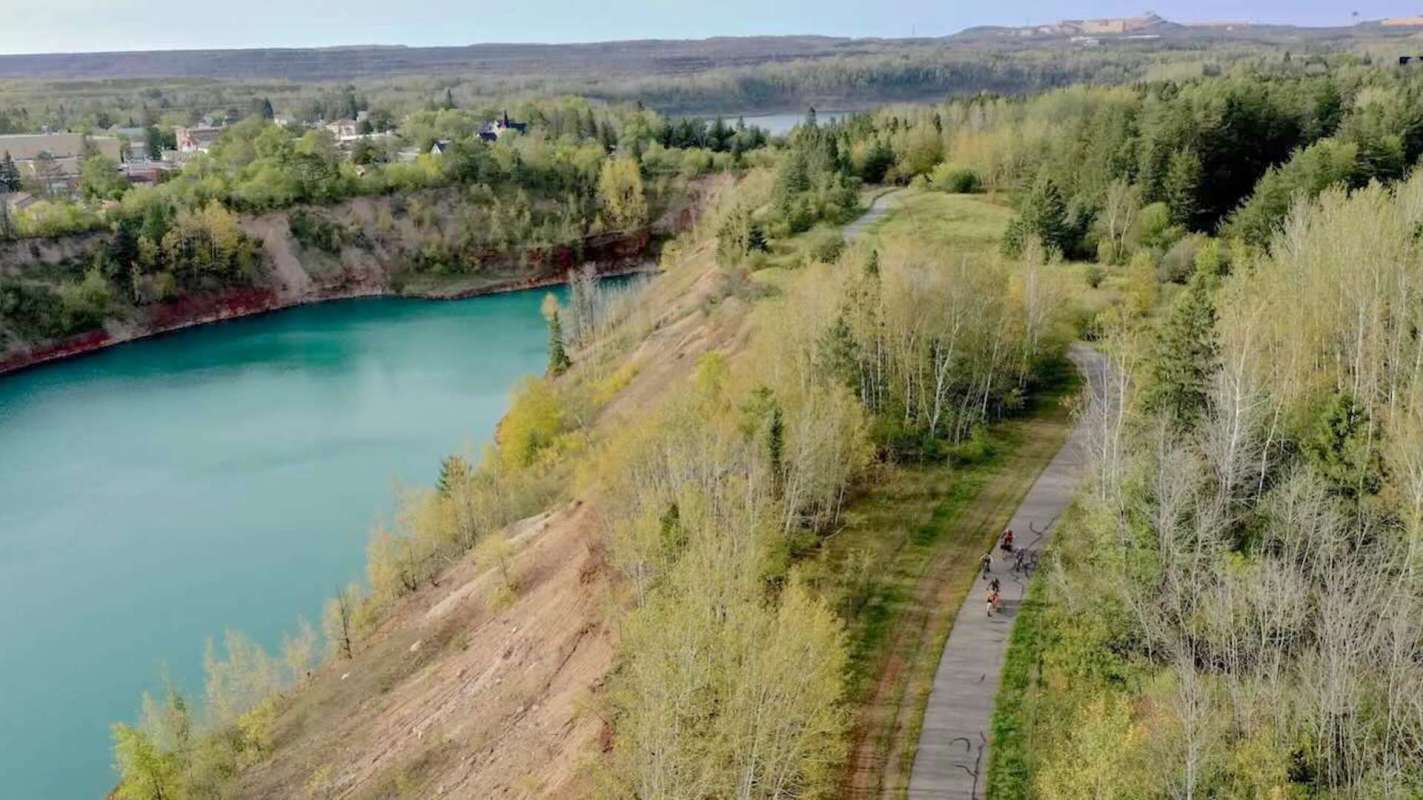 A winding path next to a blue lake and green trees with cyclists riding along the trail.