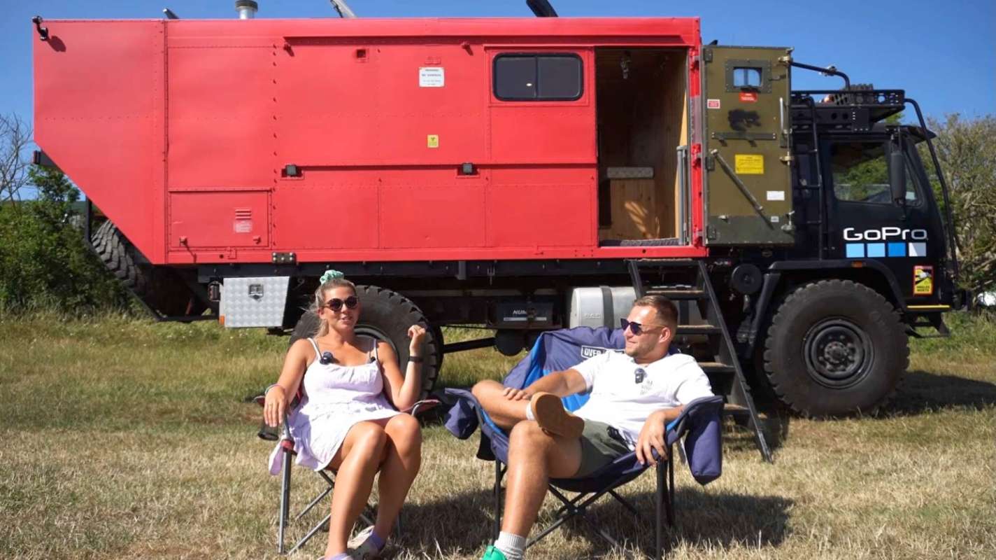 A man and a woman sitting in folding chairs near a large red vehicle in a grassy field.