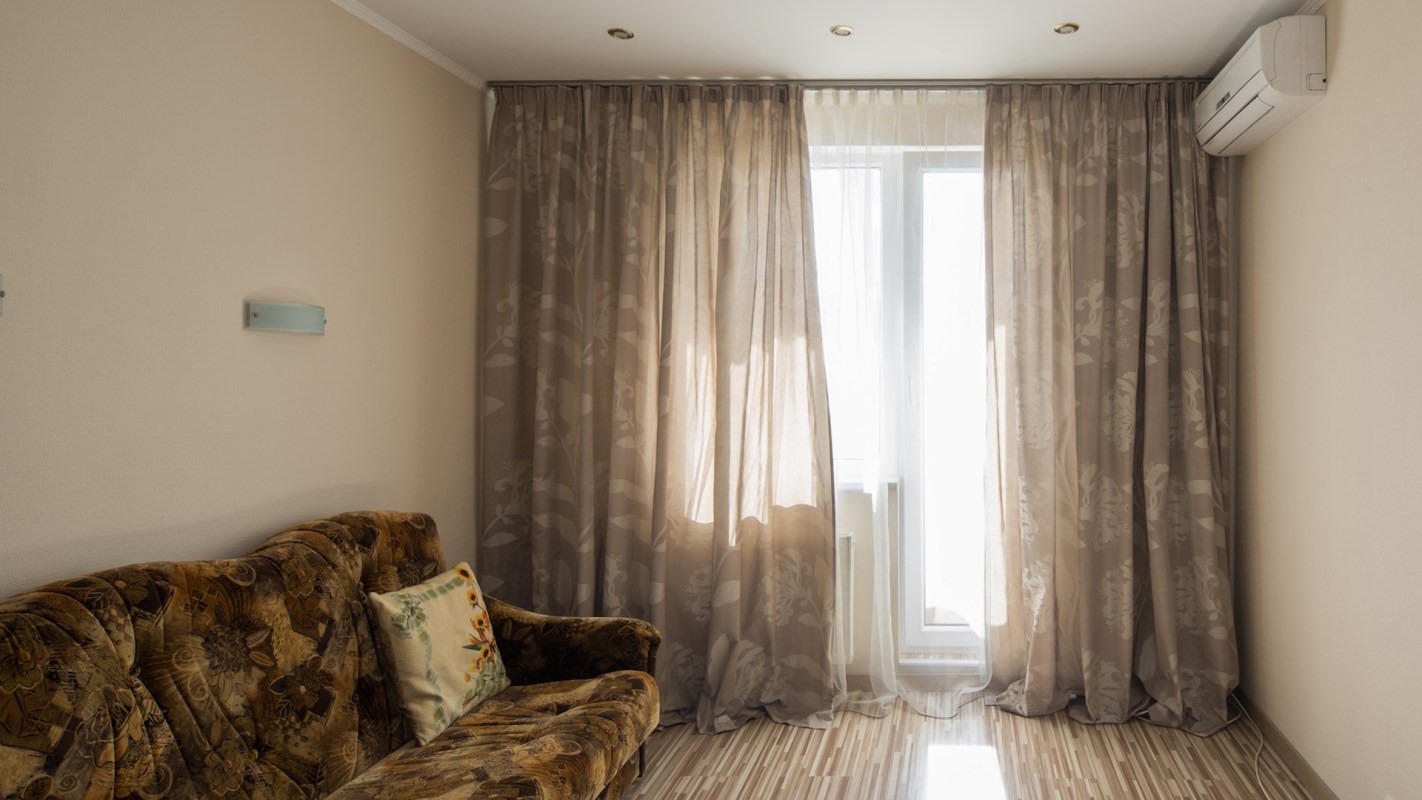 A living room with a patterned sofa, decorative pillow, and sheer curtains over a window.
