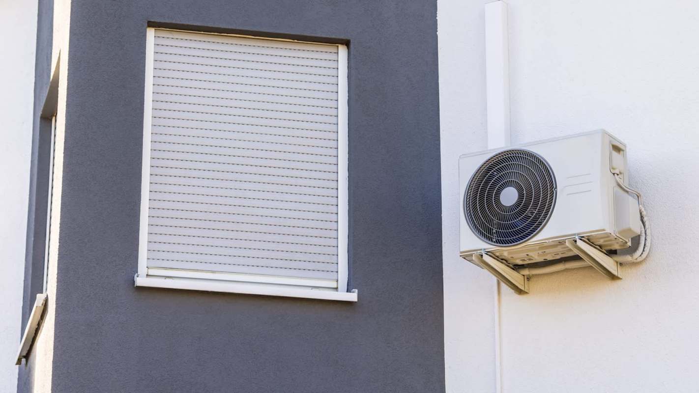 An air conditioning unit mounted on a wall beside a closed window with blinds.