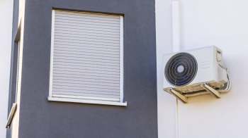 An air conditioning unit mounted on a wall beside a closed window with blinds.