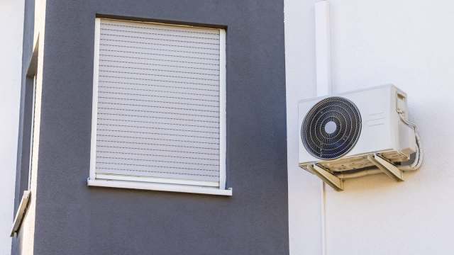 An air conditioning unit mounted on a wall beside a closed window with blinds.