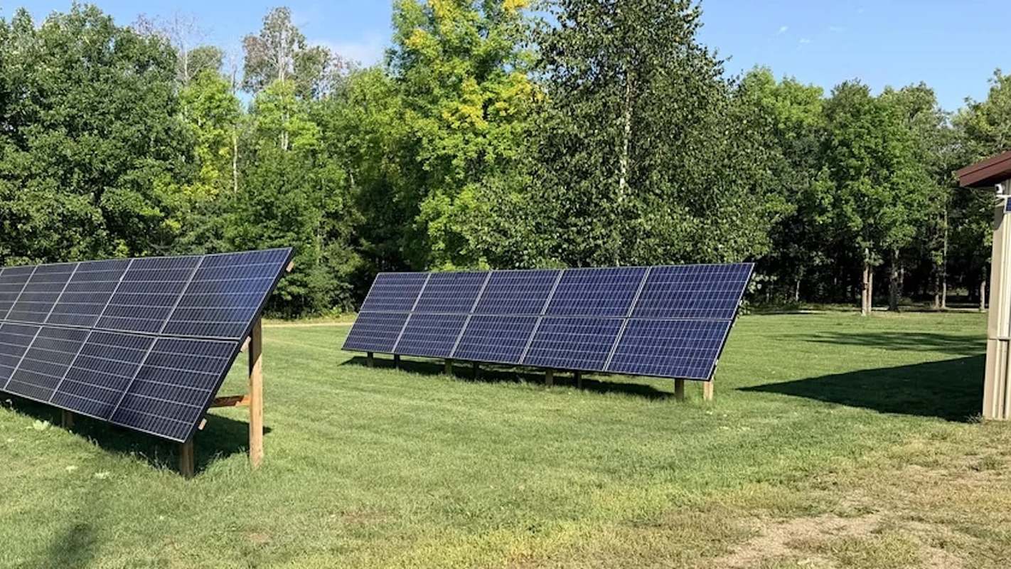 Two rows of solar panels on grass, surrounded by trees and a building.