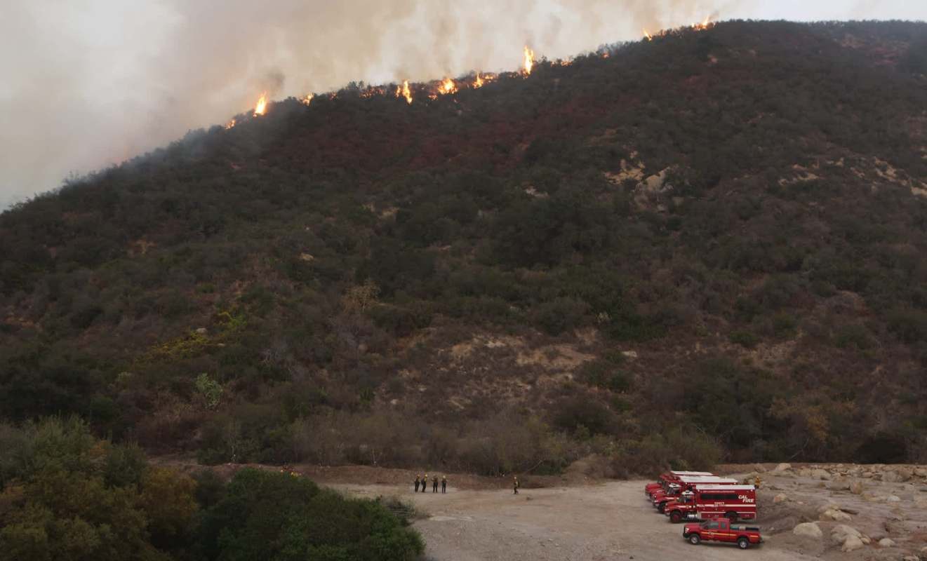 A hillside is on fire with flames and smoke, while firefighters and trucks are stationed below.