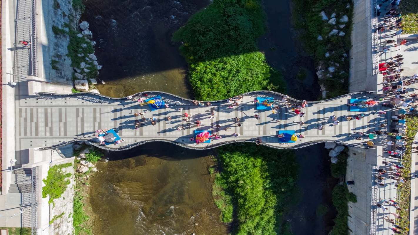 A pedestrian bridge with people walking over a river, surrounded by greenery.