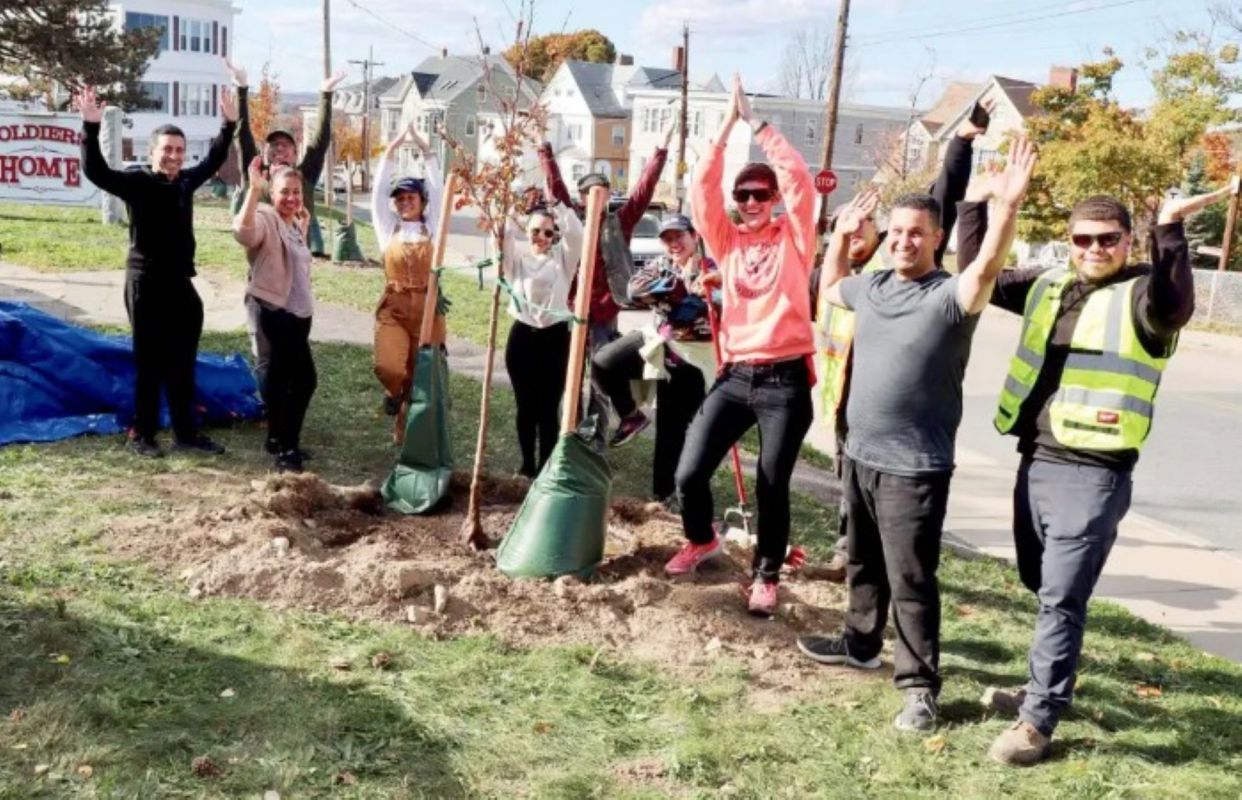 A group of people stands around newly planted trees, celebrating an outdoor planting event.