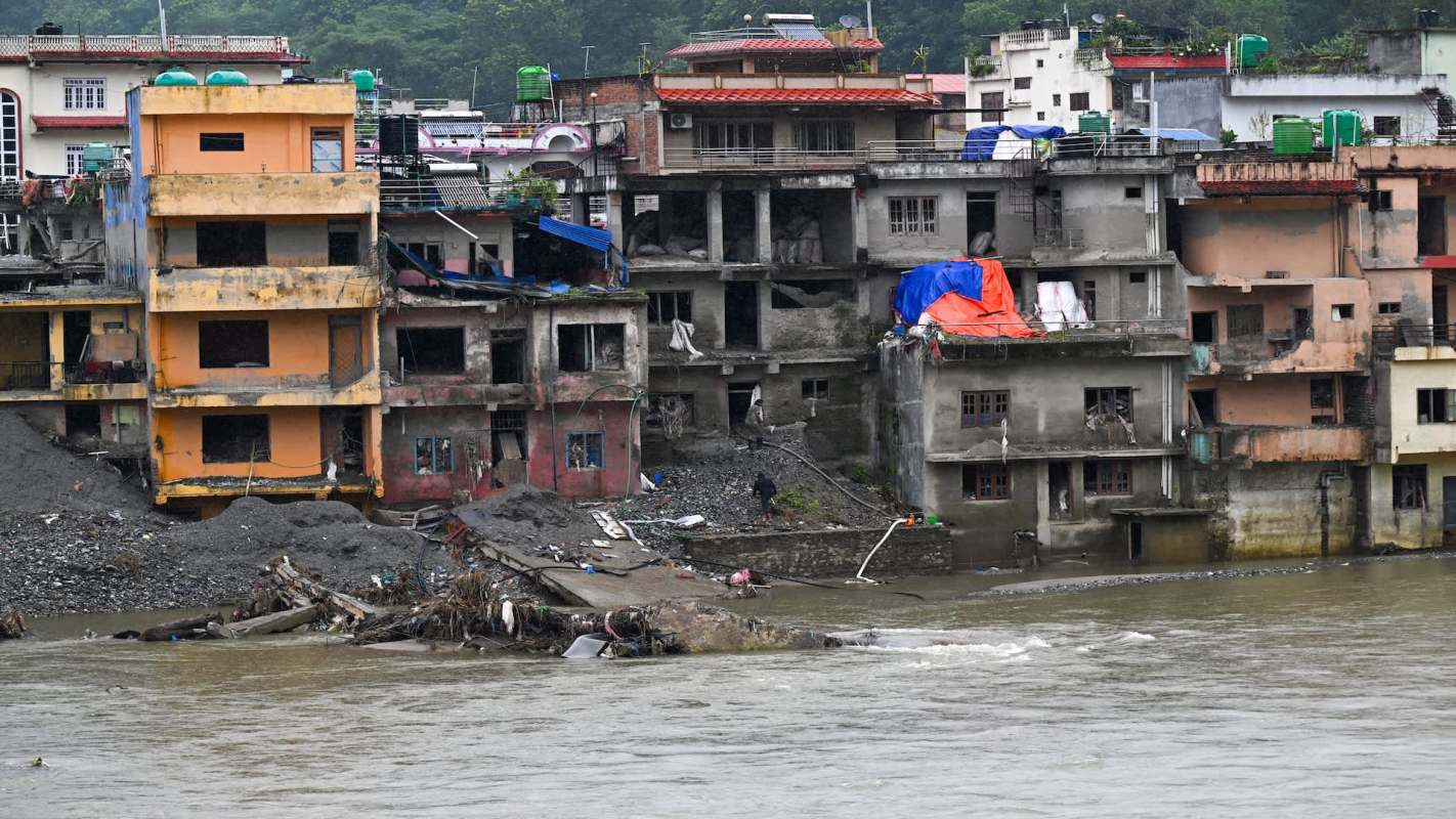 Buildings near a riverbank show signs of damage and erosion, with debris scattered along the water's edge.