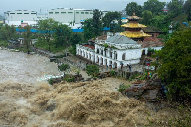 A river floods near a building, with people standing on the shore under umbrellas.