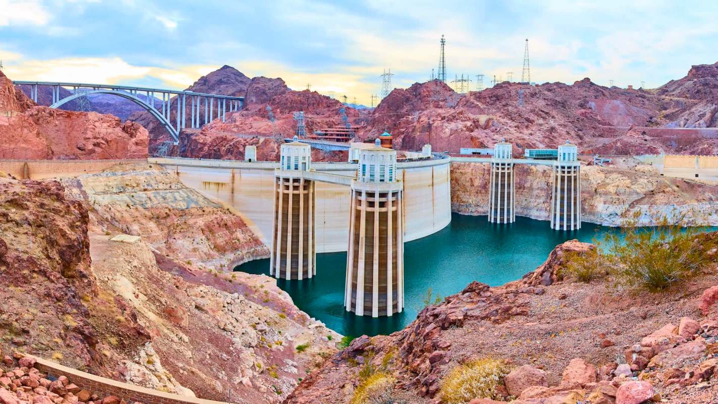 A dam with four towers stands beside a blue reservoir, surrounded by rocky hills and power lines.