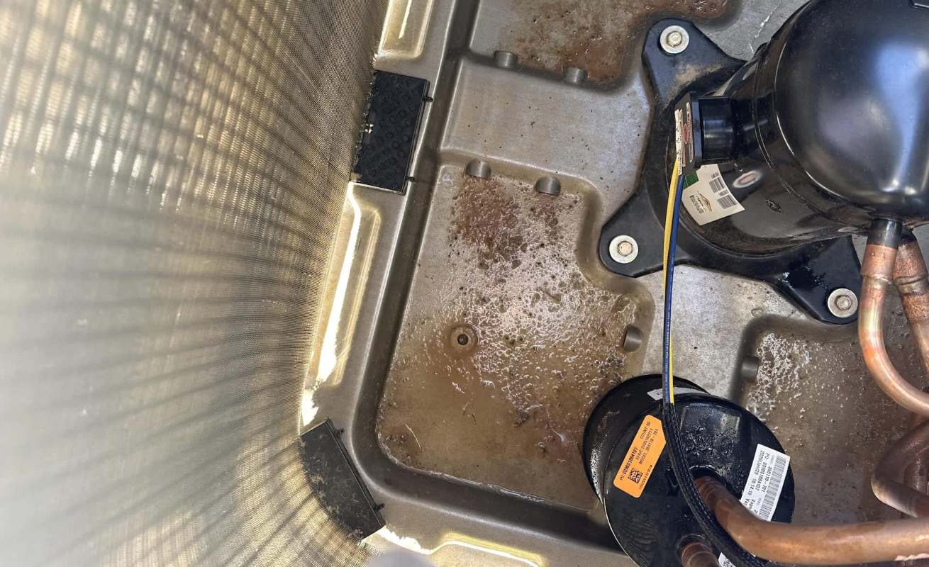 An interior view of an air conditioning unit showing a compressor, copper tubing, and a dirty, water-stained surface.