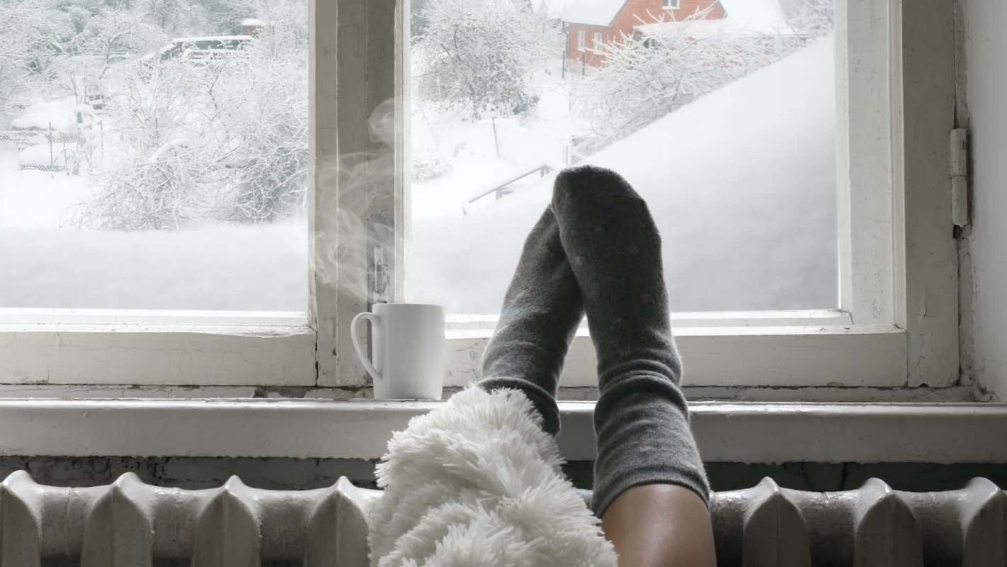 A person’s feet in gray socks rest on a windowsill next to a steaming mug, with snow-covered trees visible outside.