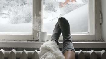 A person's feet in gray socks rest on a windowsill next to a steaming mug, with snow-covered trees visible outside.