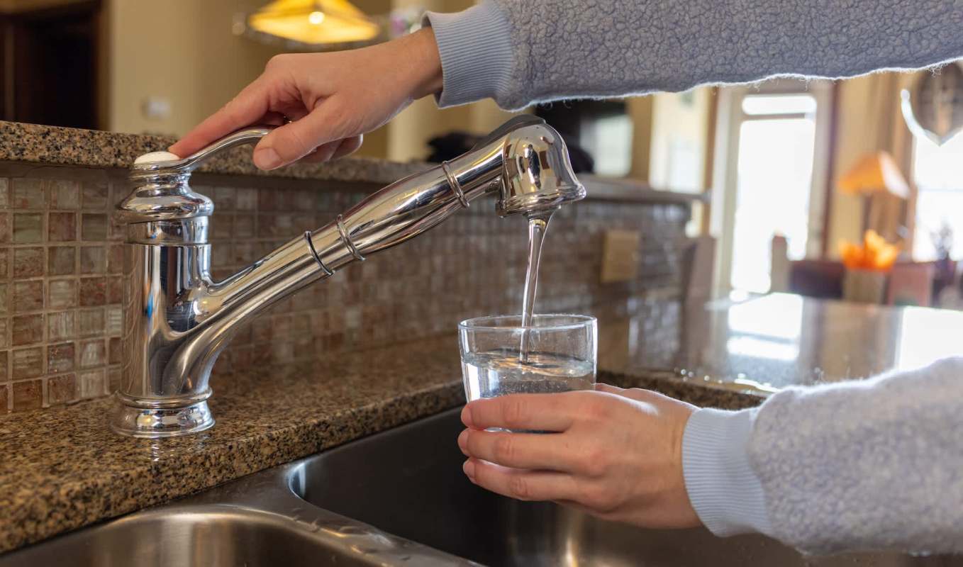 A hand fills a glass with water from a kitchen faucet.