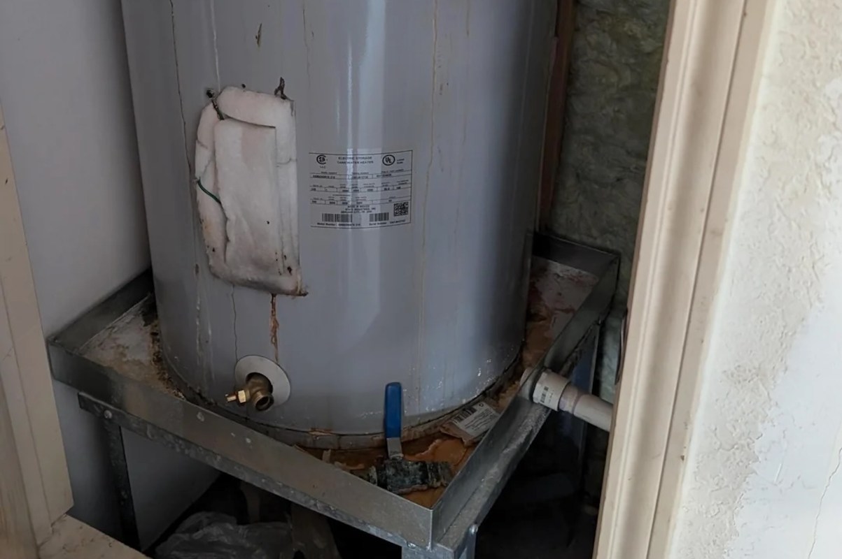 A water heater with rust and leaks placed on a metal stand in a small space.