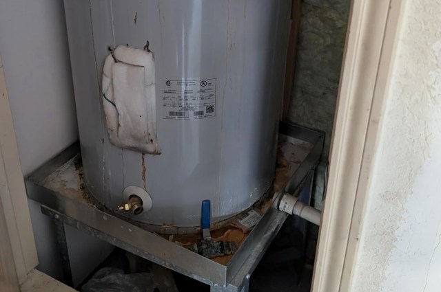 A water heater with rust and leaks placed on a metal stand in a small space.