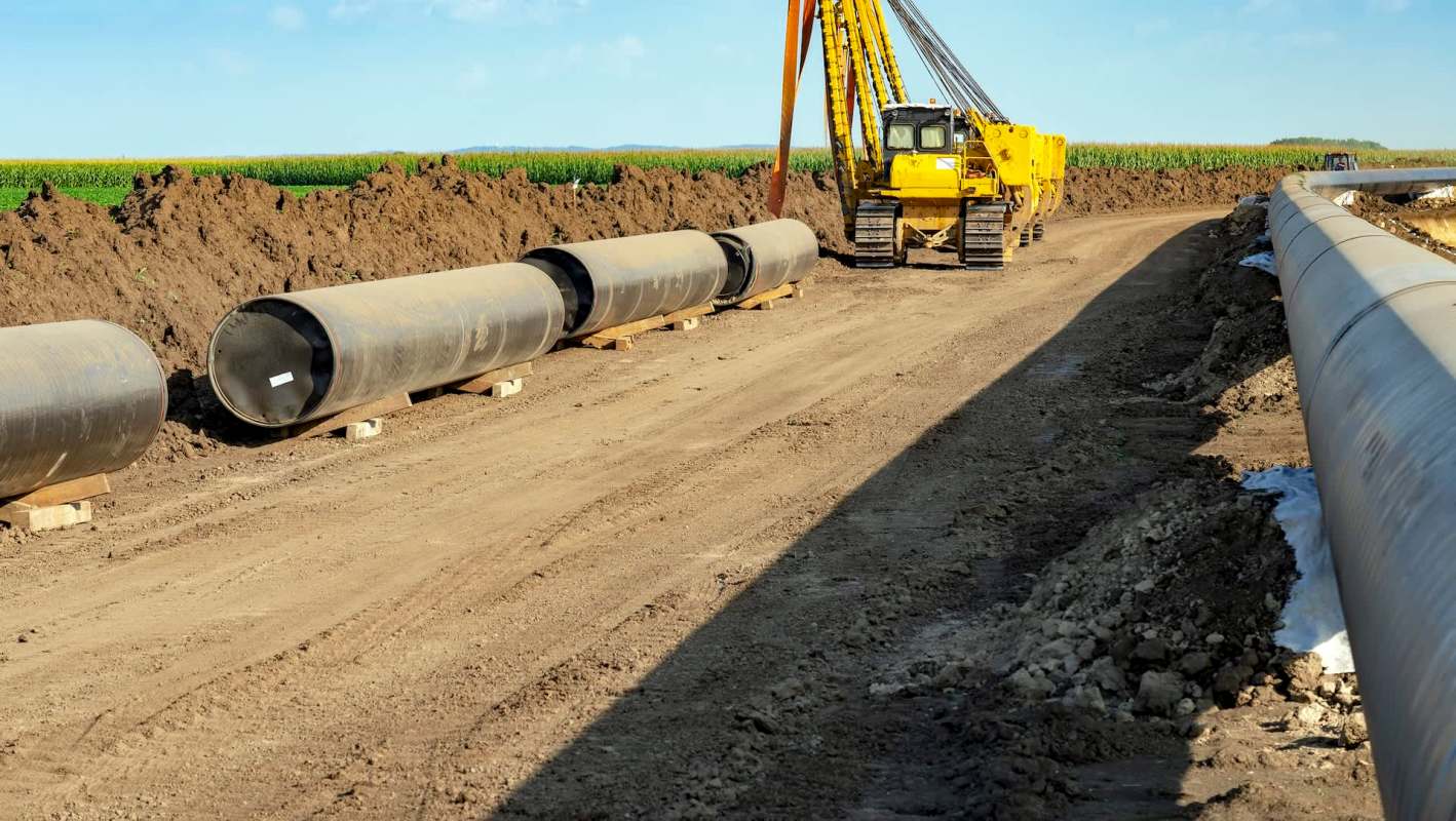 Heavy machinery is working on a construction site with large pipes lined along a dirt path near agricultural fields.