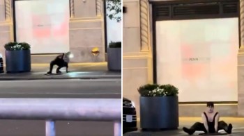 A person sitting on the street after emerging from a manhole in front of a building with large planters and a blank storefront.