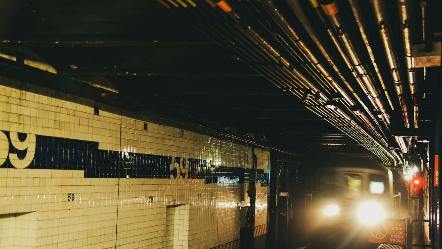 A New York City subway train approaches a platform with overhead pipes and tiled walls displaying the number 59.