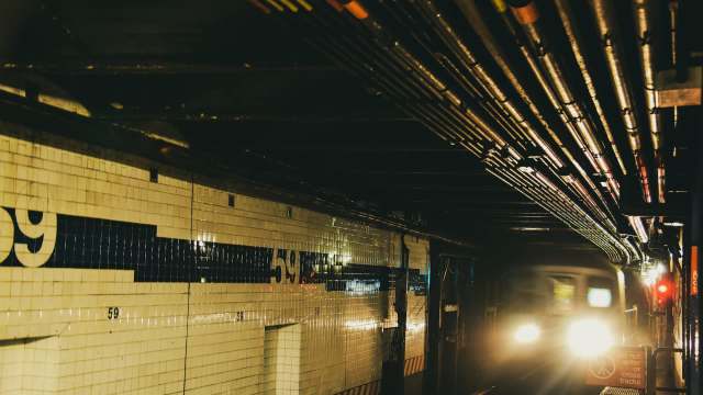 A New York City subway train approaches a platform with overhead pipes and tiled walls displaying the number 59.