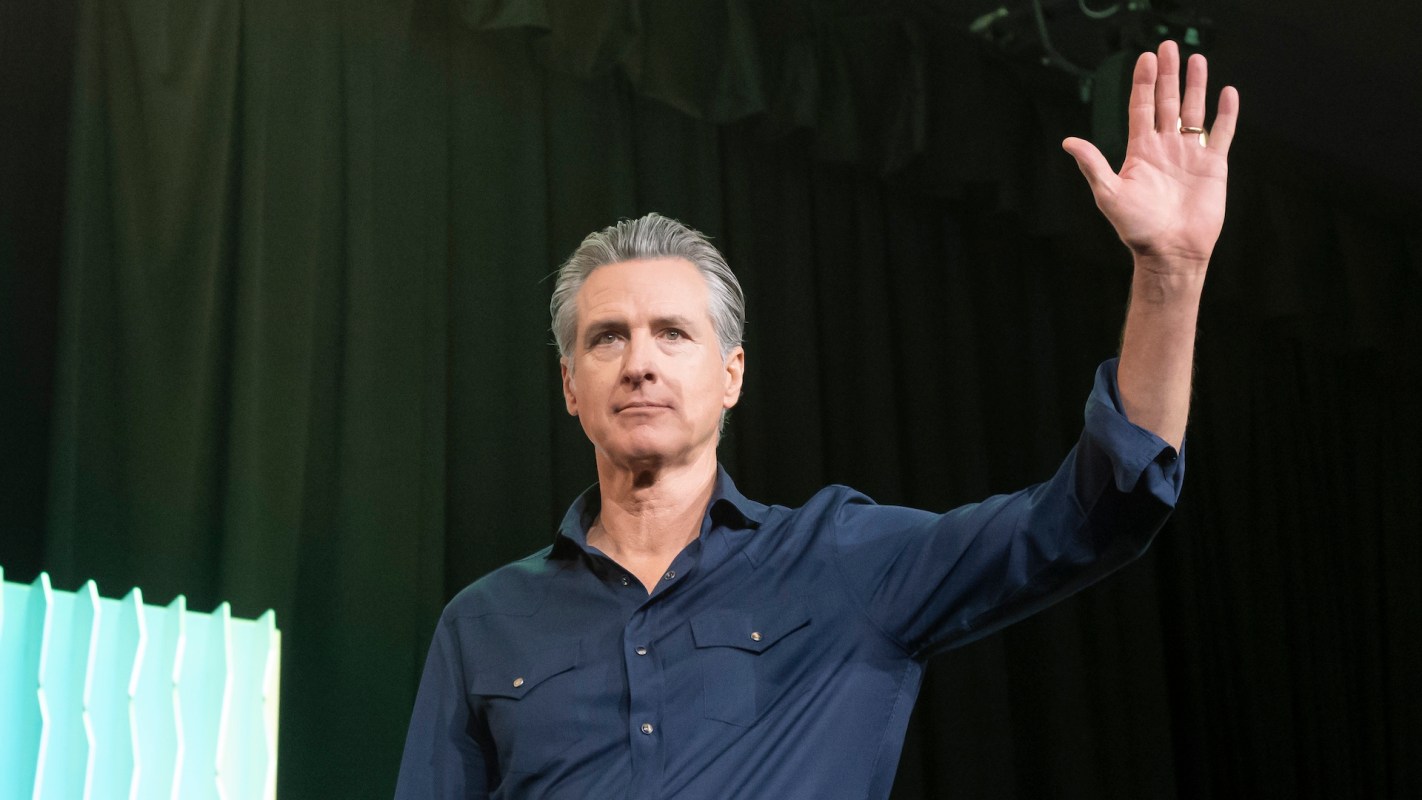 California governor Gavin Newsom raises his hand while speaking on stage with a dark backdrop.