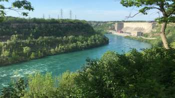 A river flows through a lush green landscape with a dam and power lines in the background.