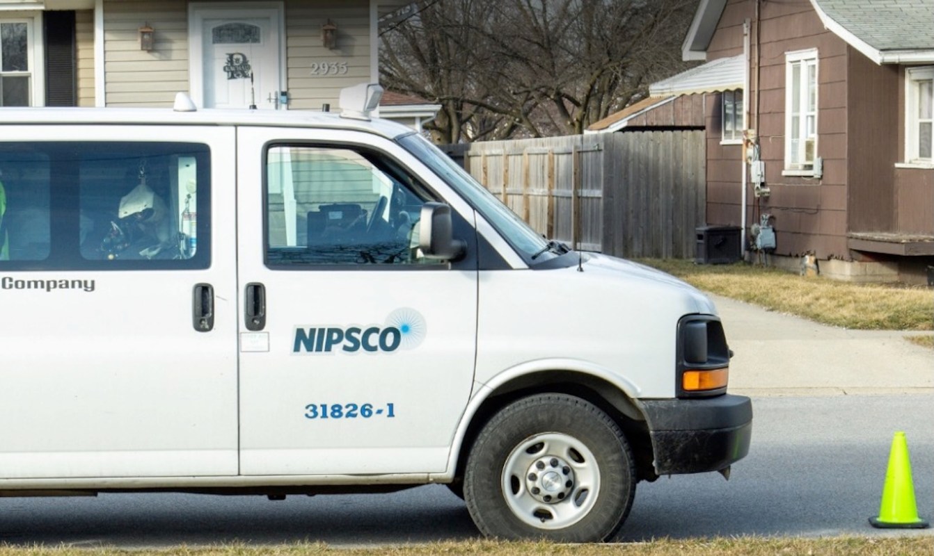 A NIPSCO service van parked on the street near a residential home with a traffic cone beside it.