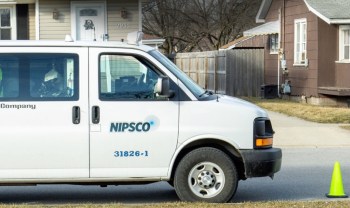 A NIPSCO service van parked on the street near a residential home with a traffic cone beside it.