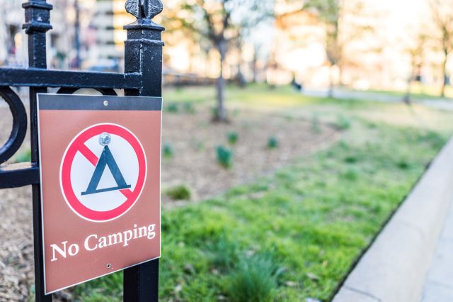 A sign on a fence displaying a "No Camping" message in a park setting.
