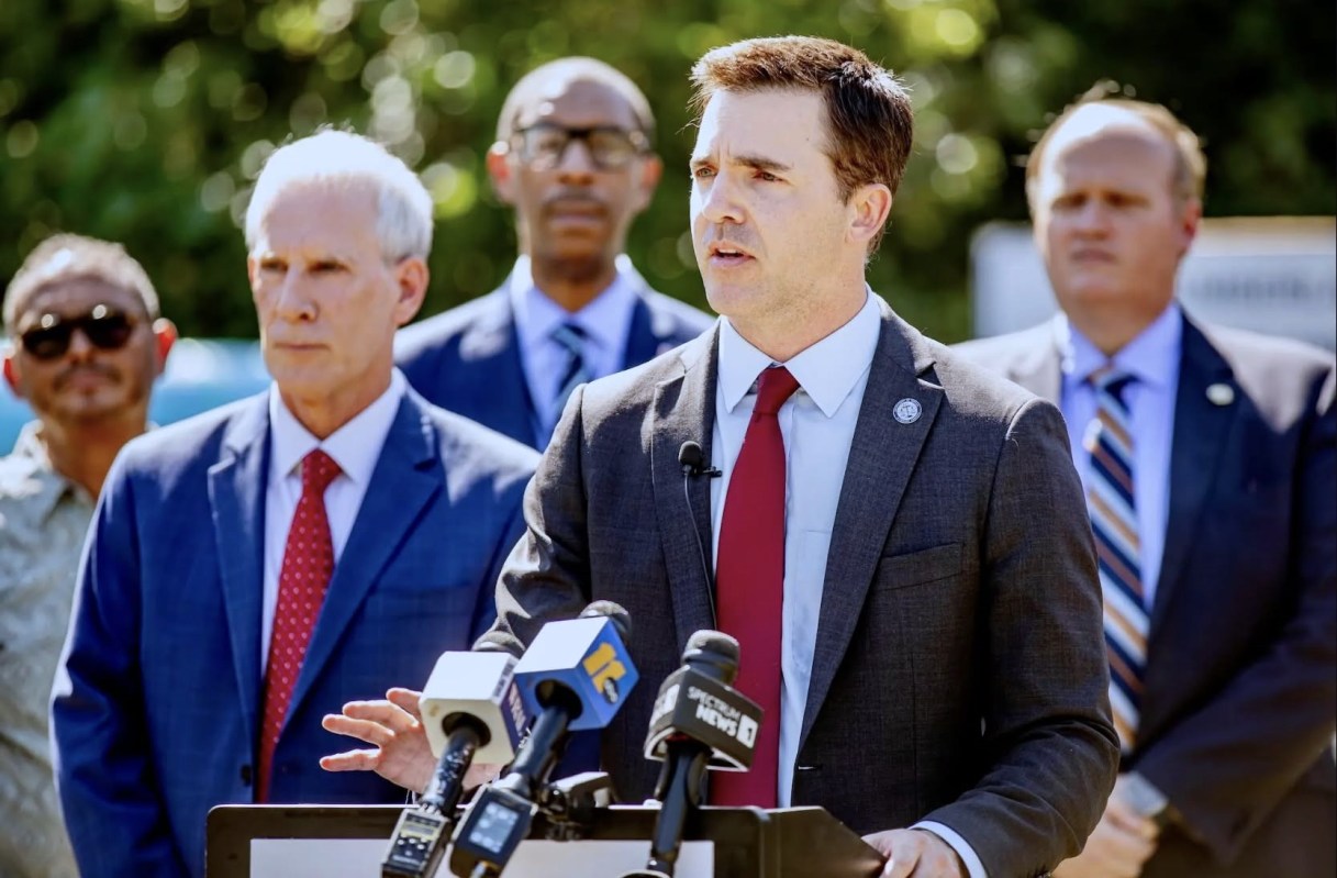 A man speaks at a podium with microphones while several people listen in the background.