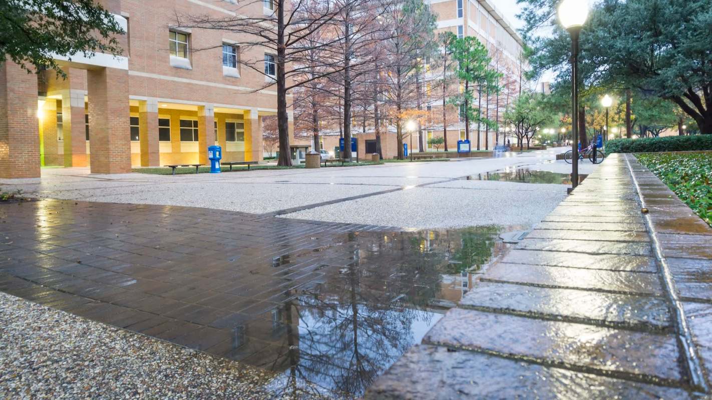 A campus courtyard with wet pavement, benches, and trees, reflecting surrounding buildings.