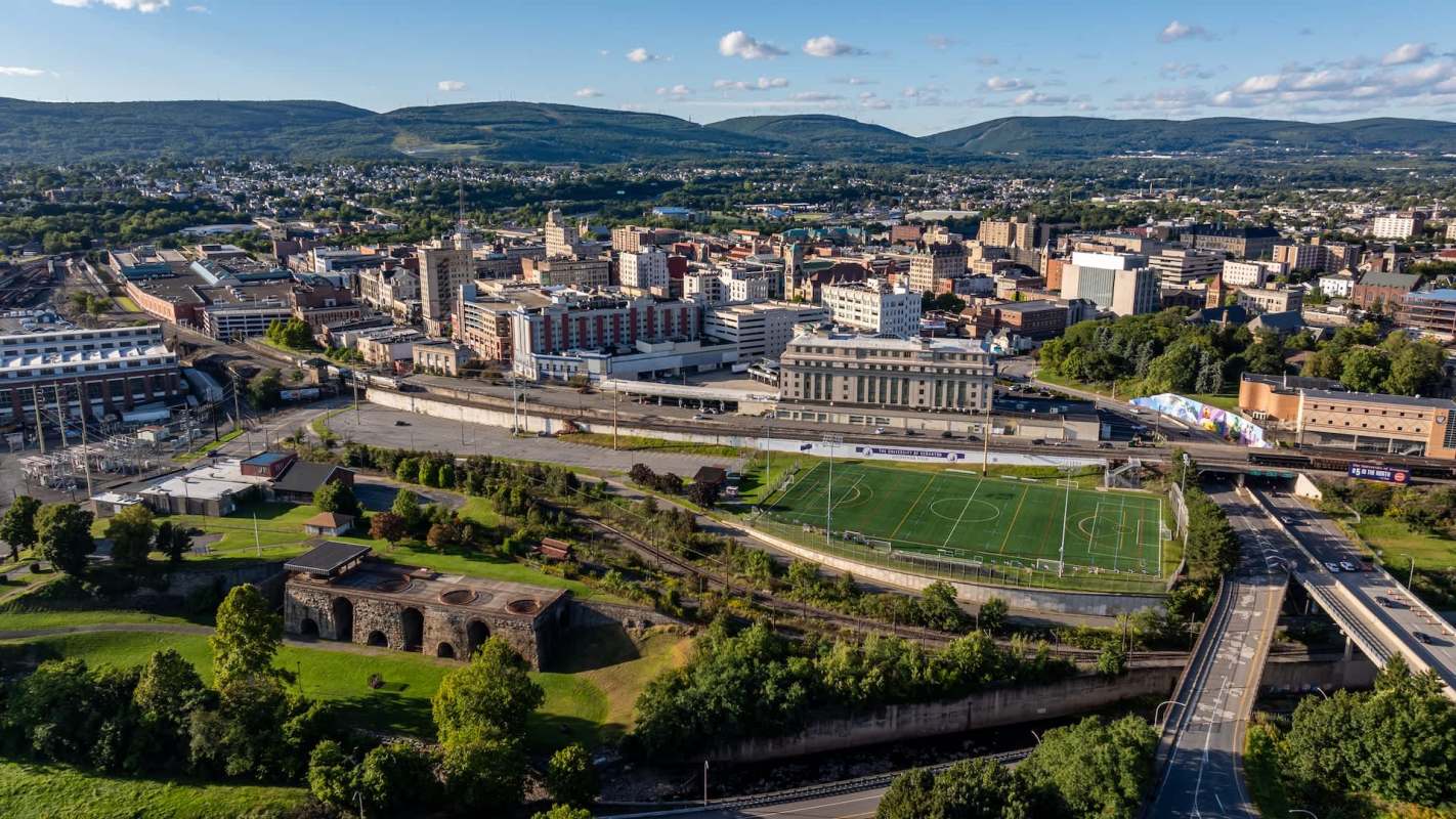 An aerial view of a city with buildings, a sports field, and surrounding greenery.