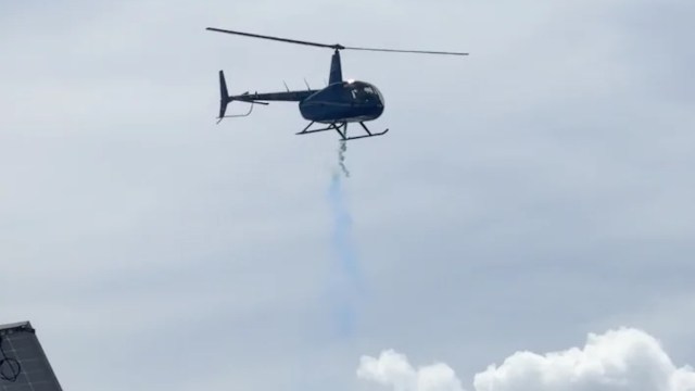 A helicopter emits a trail of blue smoke as it hovers in the sky.