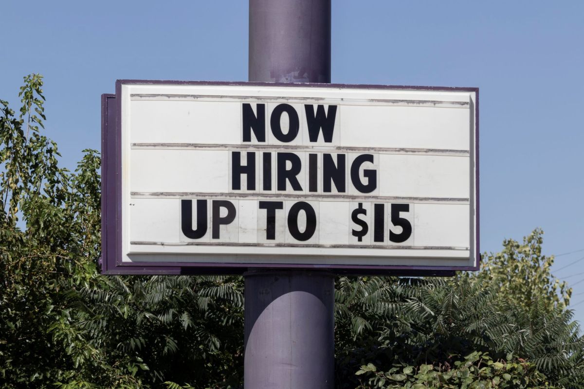 A sign that reads "NOW HIRING UP TO $15" against a clear blue sky and greenery.