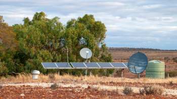 Solar panels, a satellite dish, and a water tank near a grove of trees in a dry landscape.