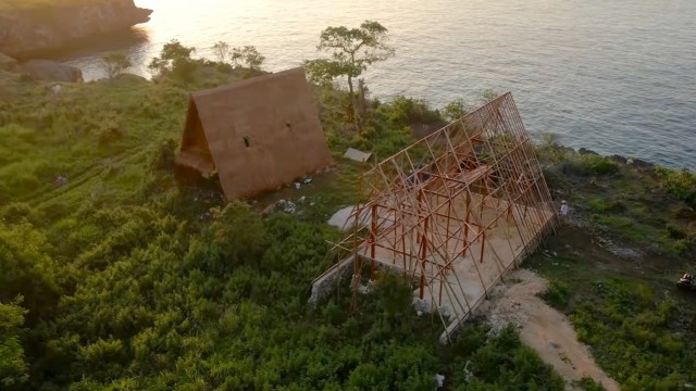 A construction site with a partially built structure and a thatched house near a coastline.
