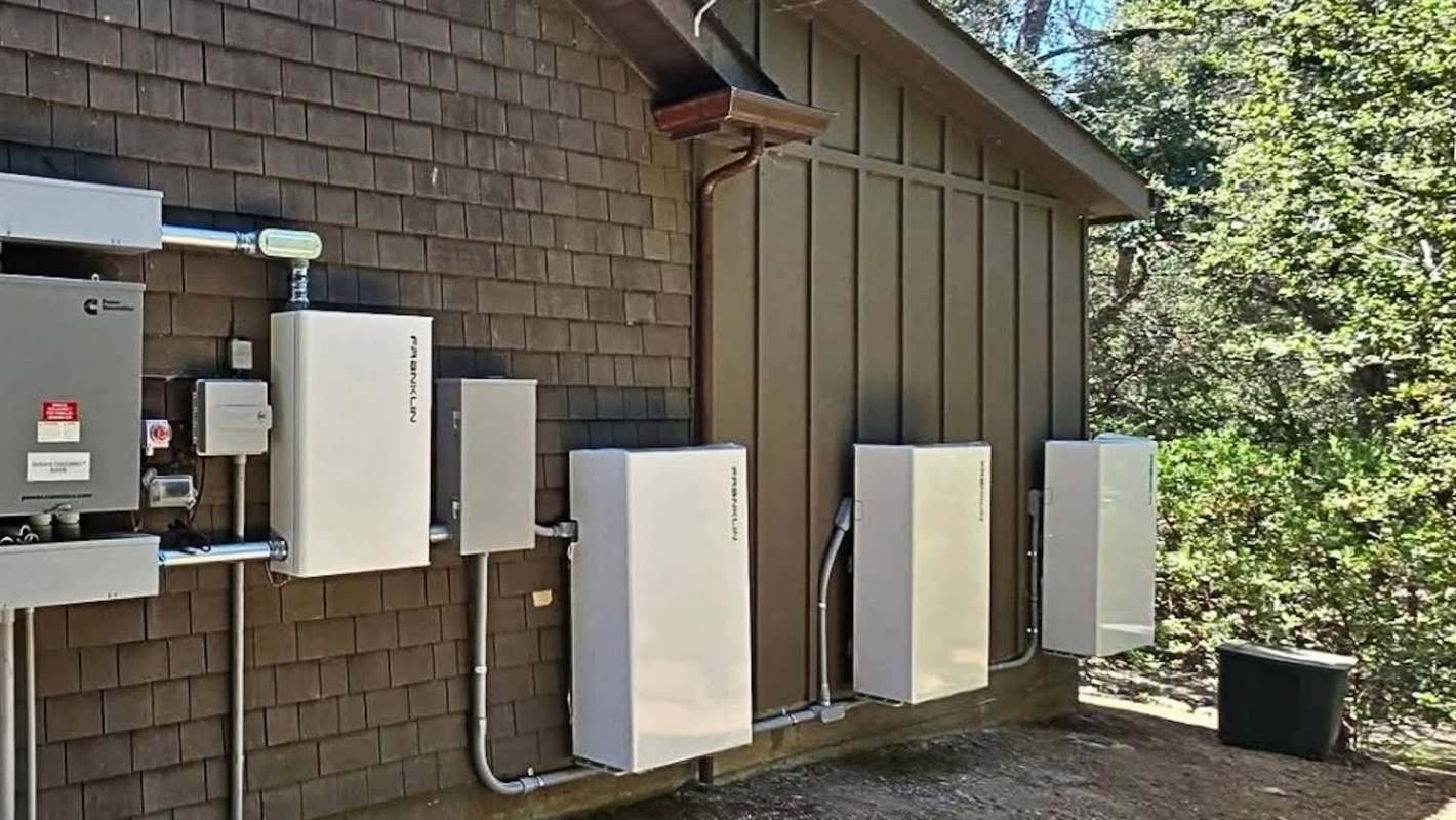 A wall-mounted setup with several white battery units and electrical components alongside a brown wooden building.